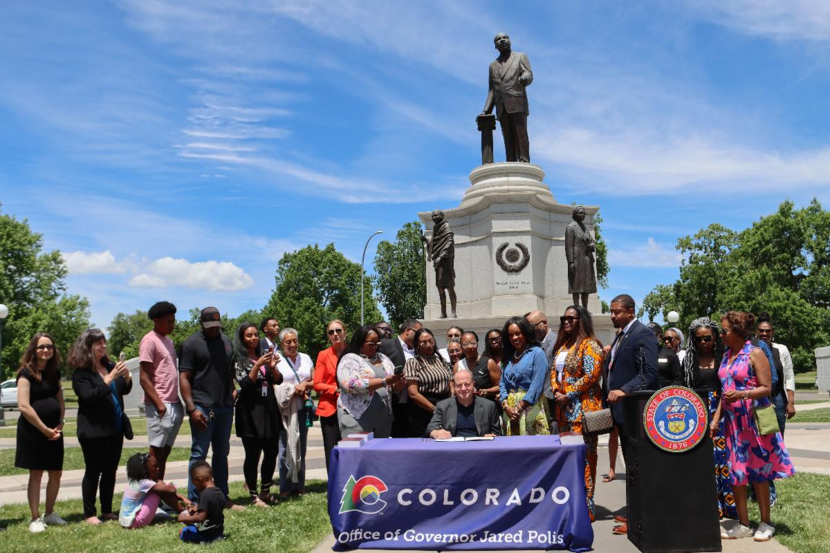 Governor Polis signs SB24-053 while seated at a table in front the Martin Luther King memorial in City Park. A crowd of advocates and legislators from the African American community surround him.