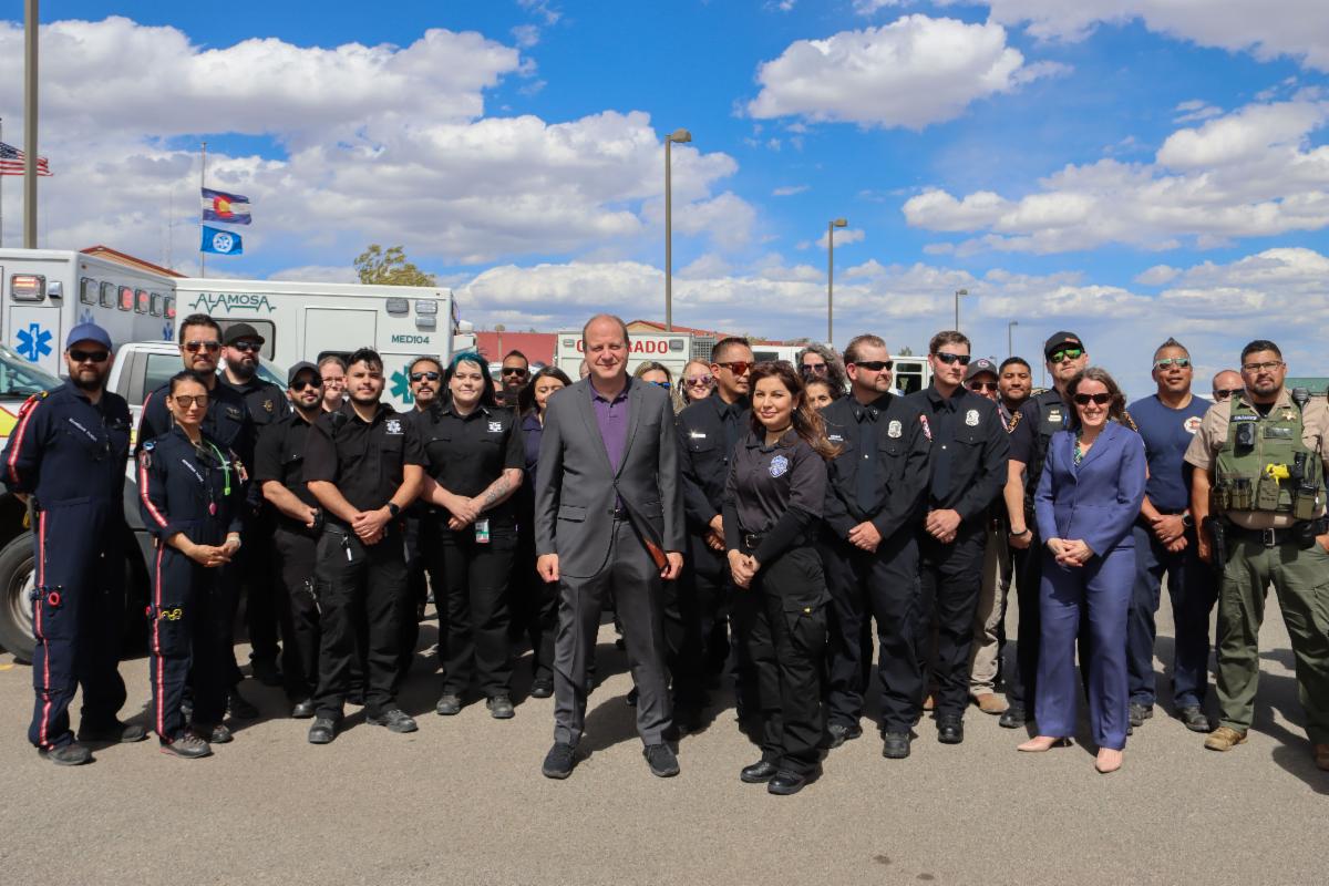 Governor Polis poses with a large group of Alamosa County first responders in front of a row of ambulances.