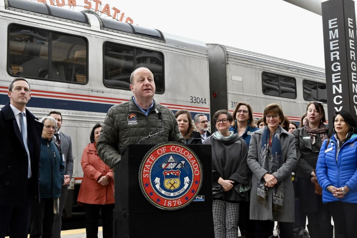 Governor Polis speaking at a podium in front of an Amtrak train at Union Station