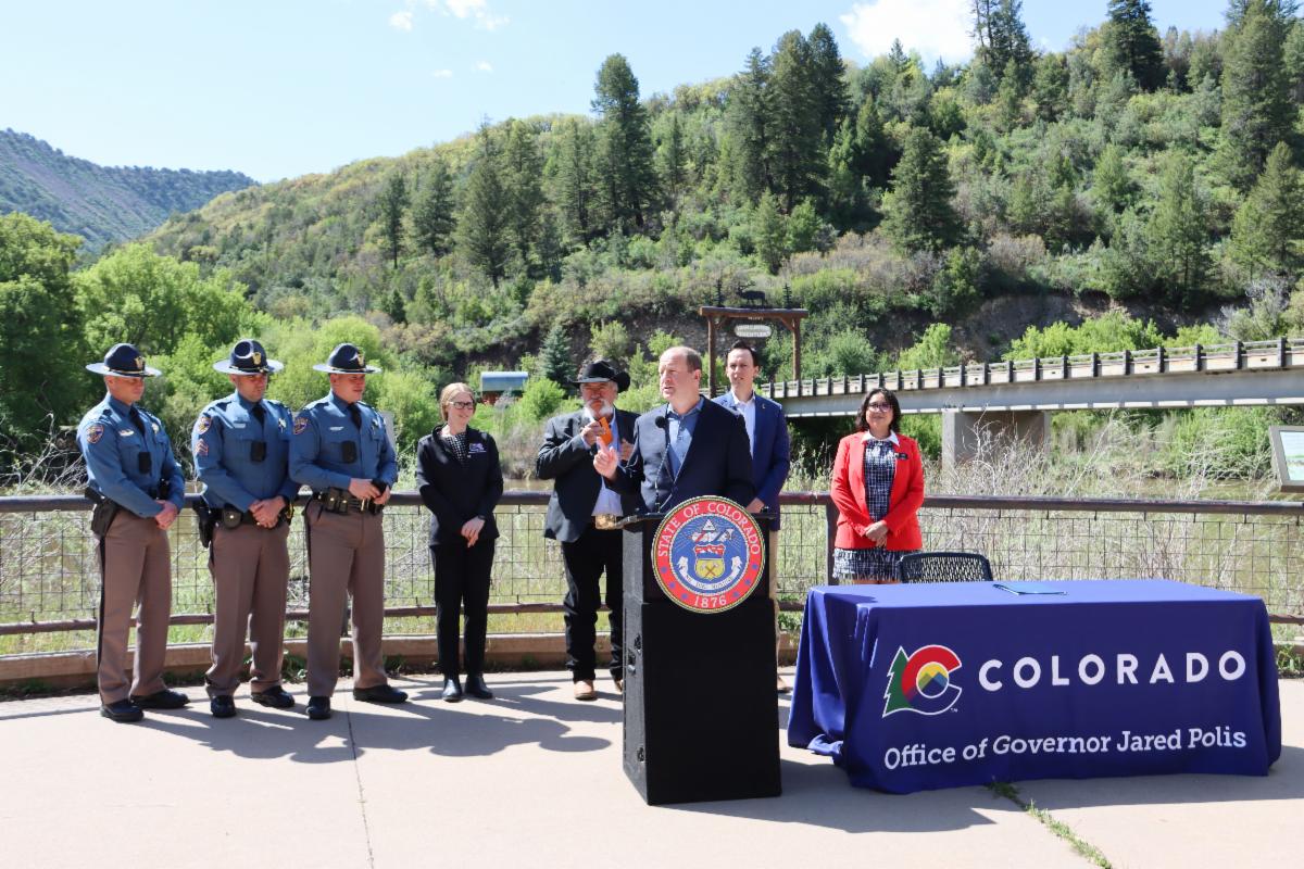 Governor Polis speaks at a podium with Senators Will and Roberts and Representative Velasco. State Patrol and CDOT Executive Director Shoshana Lew are also present.  