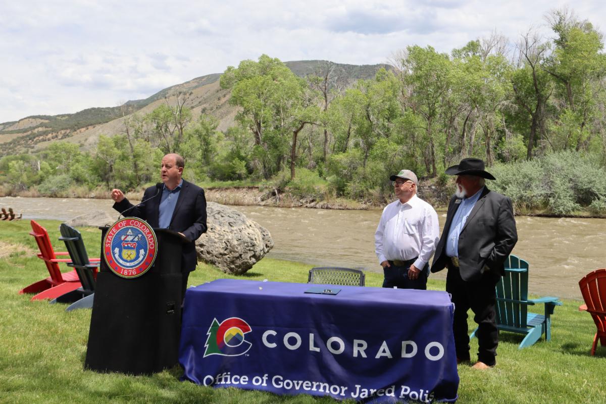 Governor Polis speaks at a podium with Senators Will and Caitlin behind him. 
