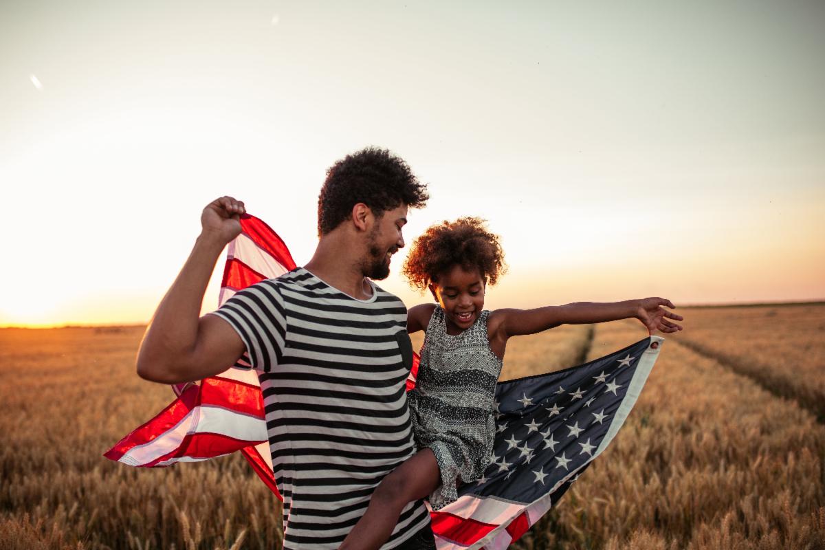 Father and daughter smiling in a field at sunset with an American flag.