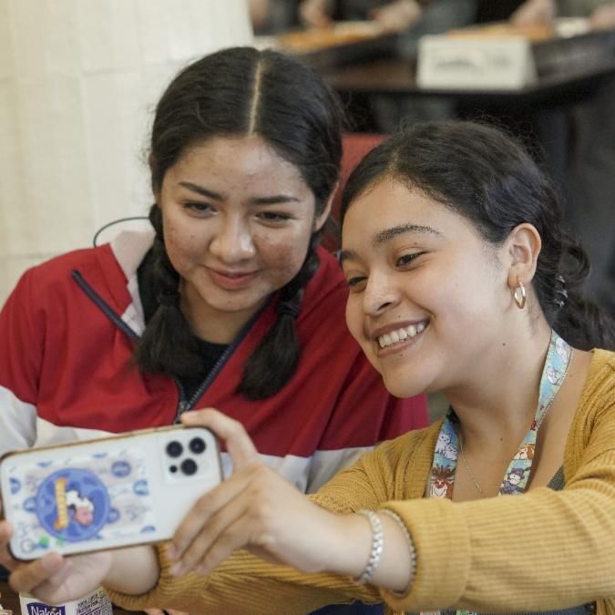 Two UC Davis students smile as they take a selfie together. One wears a red white and navy jacket. The other wears a yellow sweater and a colorful lanyard.