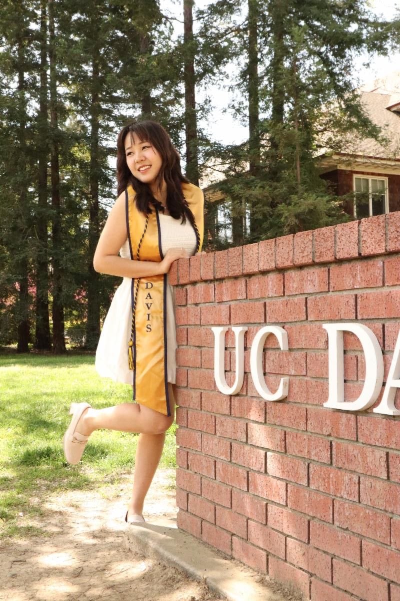 Image of a UC Davis student wearing a gold graduation stole in front of a brick monument displaying UC Davis