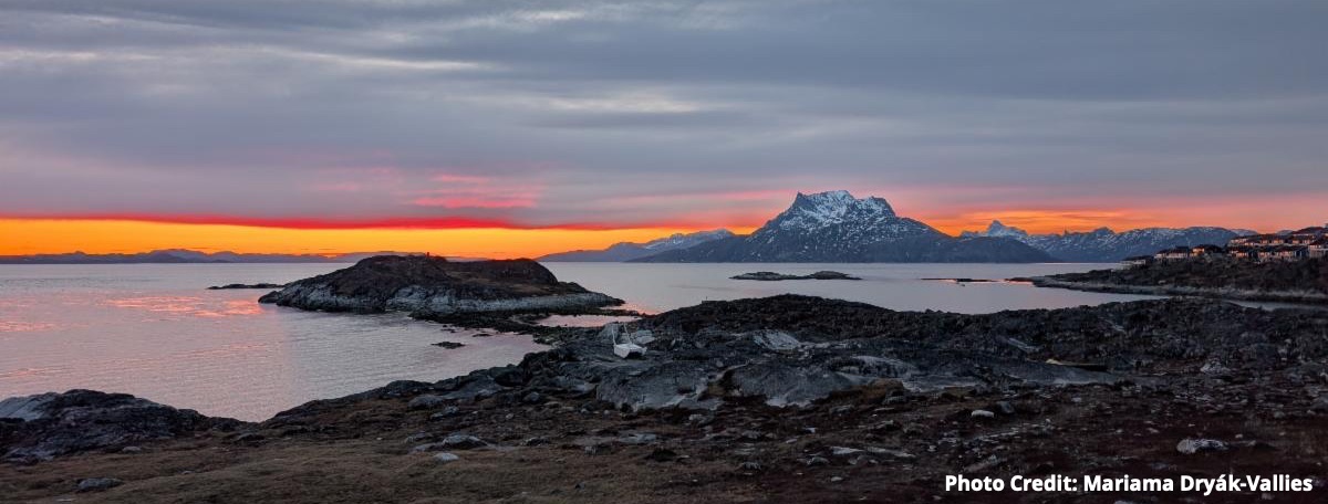 Photo of a 3am sunrise over the fjord - colors in the sky range from a cool, dark blue to highlighter orange and pink. A mountainous, snow-covered island is visible in the distance. Photo by Mariama Dryák-Vallies