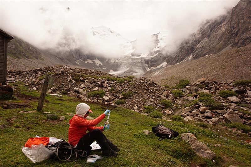 Person wearing a red jacket sitting on grass in a mountainous area holding an object with rocky terrain and misty peaks in the background scattered gear is placed nearby