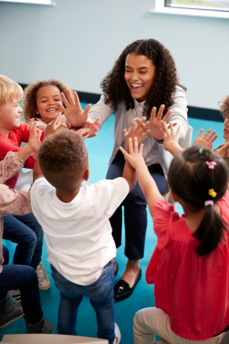 Elevated view of infant school children in a circle in the classroom giving high fives to their smiling female teacher_ vertical_ close up
