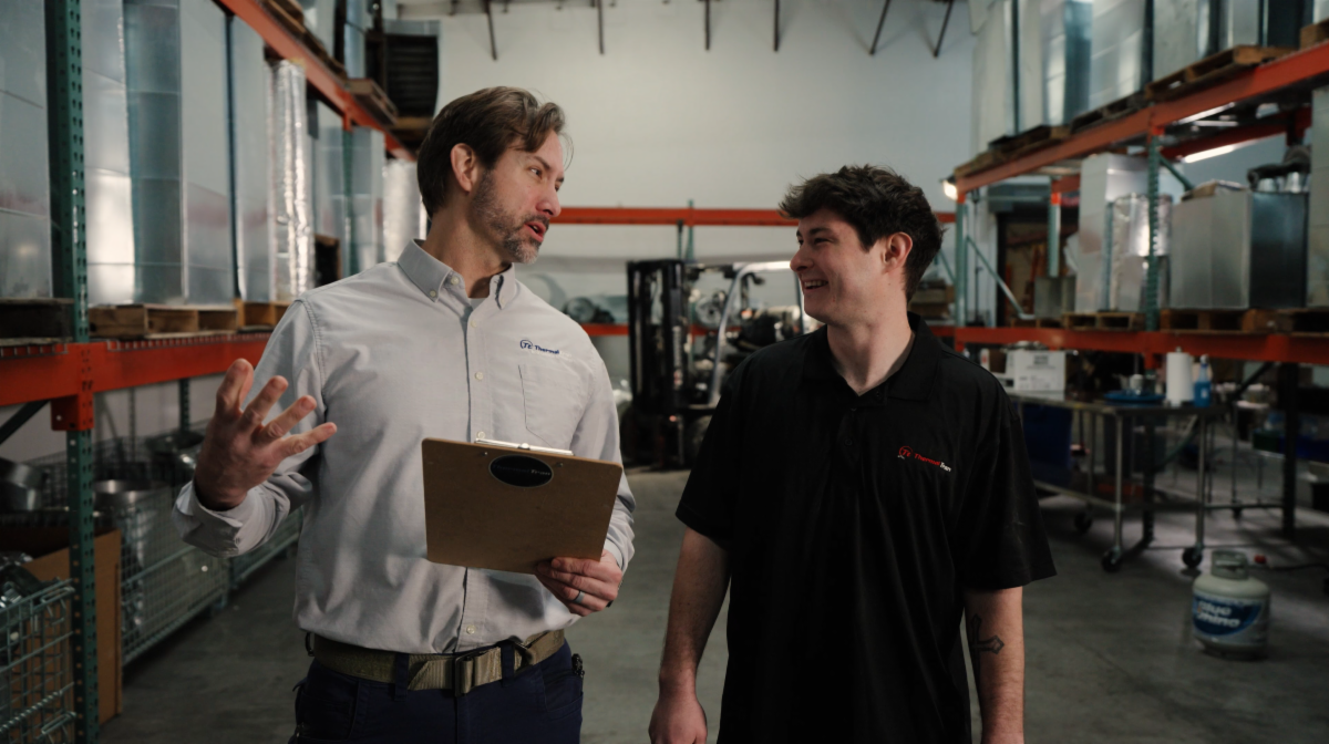 Two men stand in a warehouse aisle, talking and smiling. One holds a clipboard and gestures while speaking, and the other listens. Shelving units, equipment, and a forklift are visible in the background.
