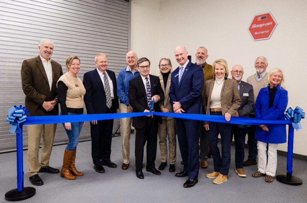 A group of community leaders and staff stand behind a large blue ribbon during a ribbon-cutting ceremony indoors. One man at the center holds scissors to cut the ribbon, while others smile. A Snap-on Multimeter sign is mounted on the wall behind them.