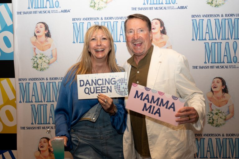 Two people smile and pose in front of a “Mamma Mia!” backdrop, holding signs that read “Dancing Queen” and “Mamma Mia” at a themed event.