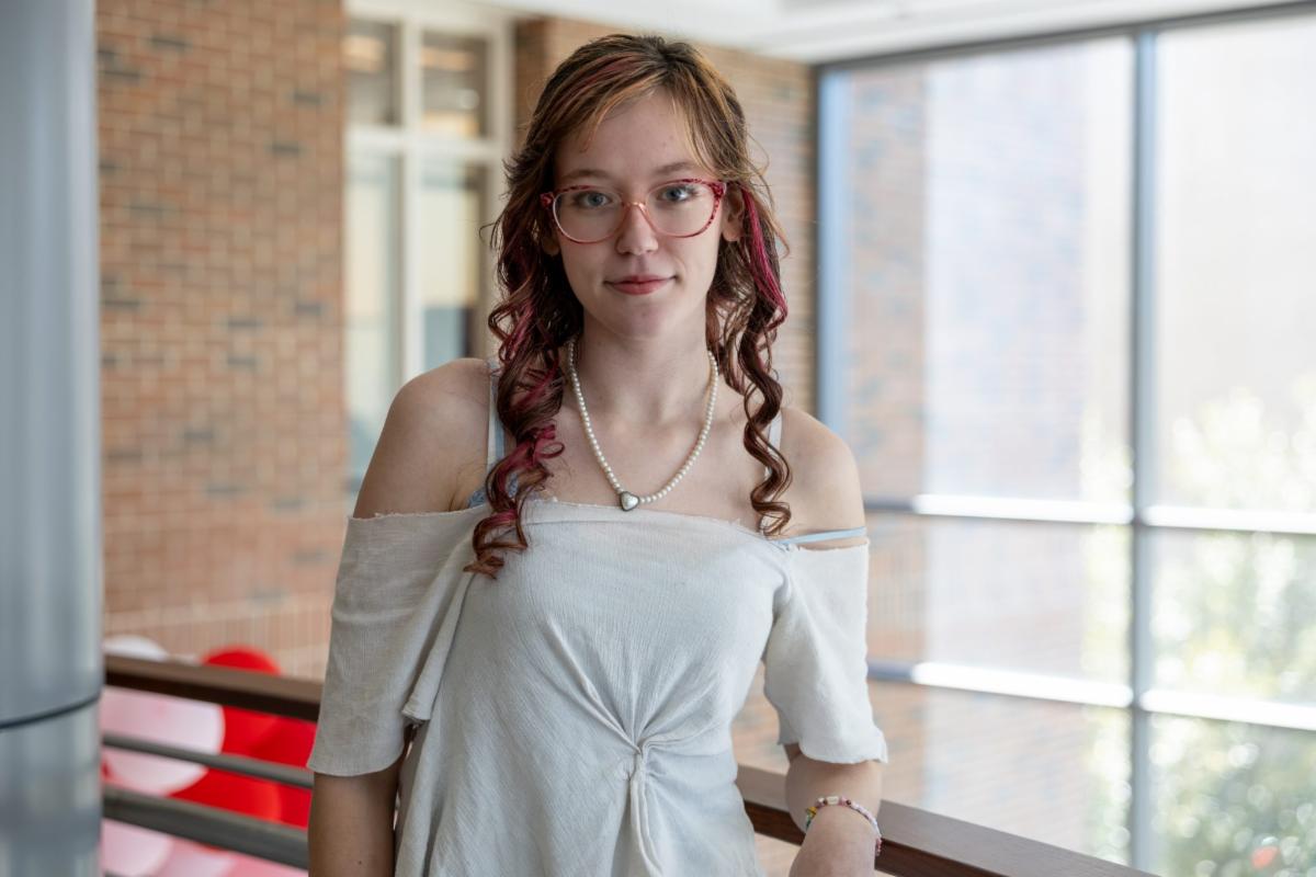 A young woman with glasses and curled hair stands indoors by a railing, wearing an off-the-shoulder top and a necklace, with large windows and a brick wall in the background.
