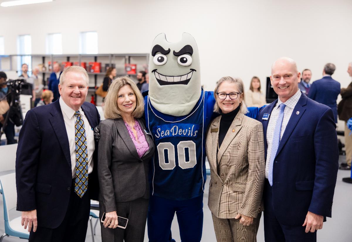 Four adults in business attire stand smiling with a Sea Devils mascot wearing a blue jersey with the number 00. The group poses together indoors at an event, with other attendees and equipment visible in the background.