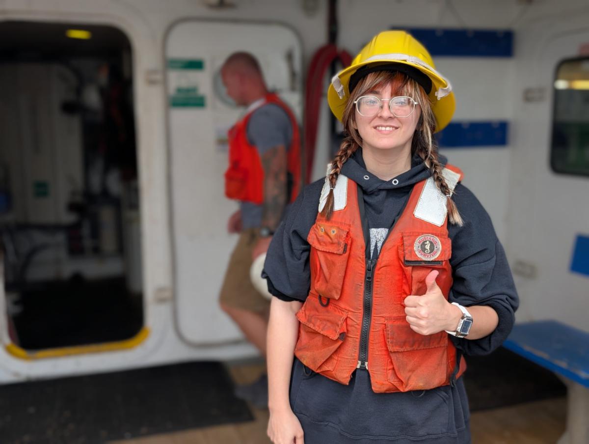 Woman wearing a yellow hard hat, clear glasses, and an orange safety vest over a dark hoodie gives a thumbs-up while standing on what appears to be a boat deck. A second person in a red vest is blurred in the background near an open doorway.