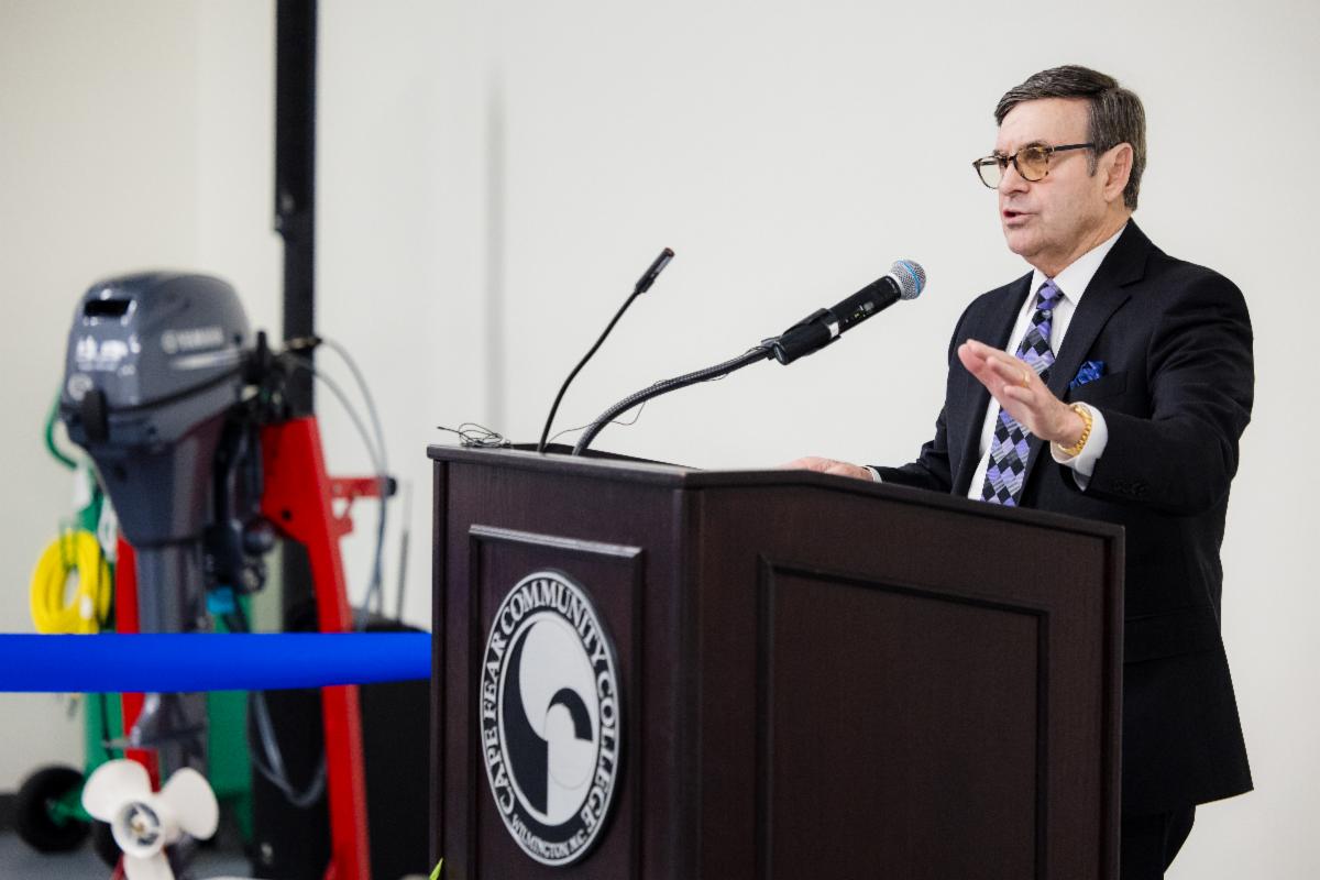 A man in a dark suit and patterned tie speaks at a wooden podium with the Cape Fear Community College seal on the front. He gestures with one hand while addressing an audience. A microphone is positioned in front of him, and marine equipment is visible in the background.