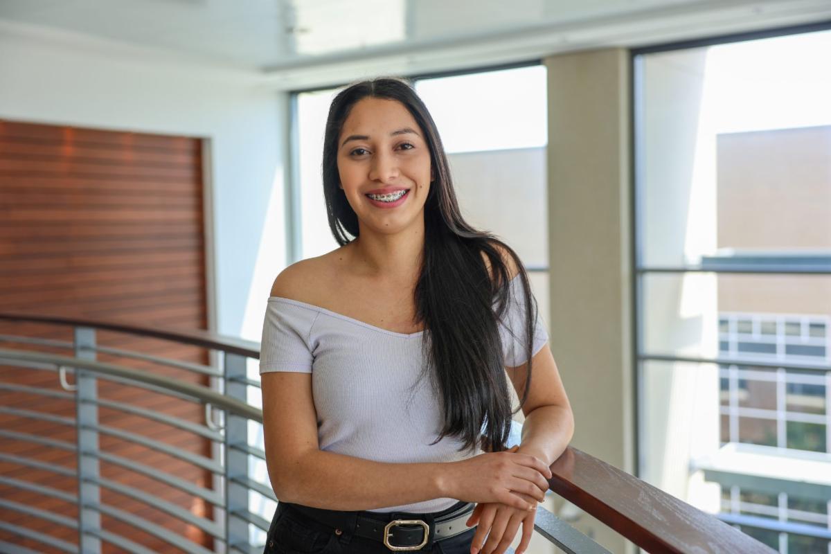 Smiling young woman with long dark hair and braces standing indoors by a railing in a bright modern building with large windows.