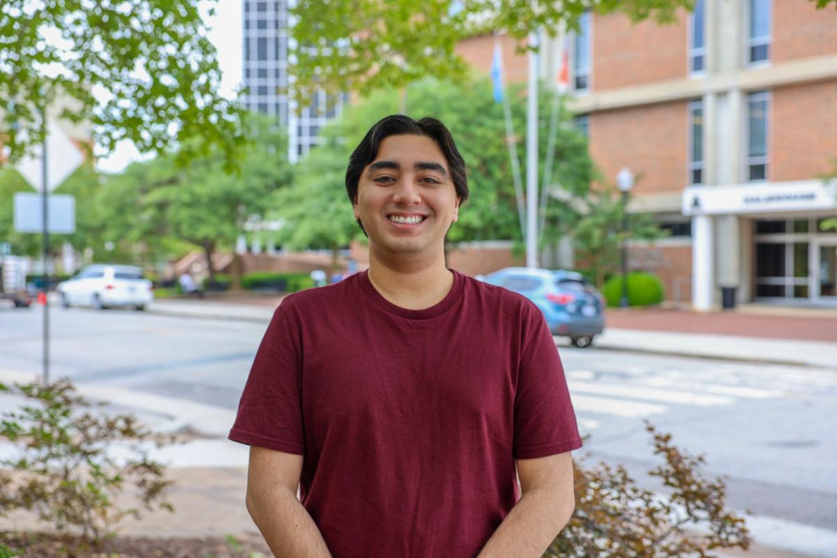 Smiling young man in a maroon T-shirt standing outdoors on a tree-lined city street near office buildings and parked cars.