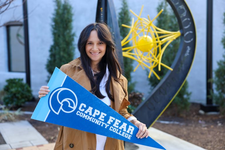 A woman standing outdoors holding a blue Cape Fear Community College pennant. A decorative sculpture with a yellow starburst design is visible behind her.