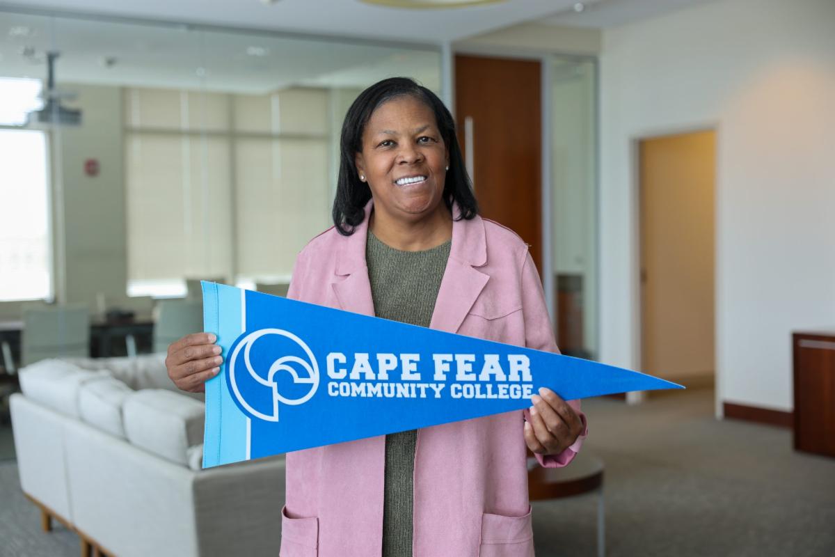 A woman stands indoors holding a blue pennant that reads “Cape Fear Community College,” smiling at the camera. She wears a pink jacket over a green top, and a modern office or lounge space with glass walls and seating is visible in the background.