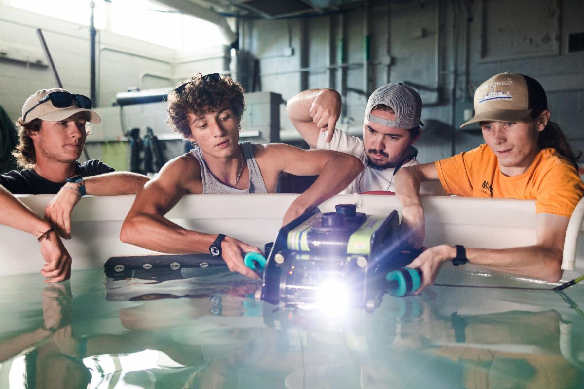 Four young men lean over the edge of a large indoor water tank, working together to operate a small underwater remotely operated vehicle (ROV) with bright front lights. One holds the device steady in the water while another points at it, and the others watch closely. The setting appears to be an industrial or workshop environment with concrete walls and overhead lighting.