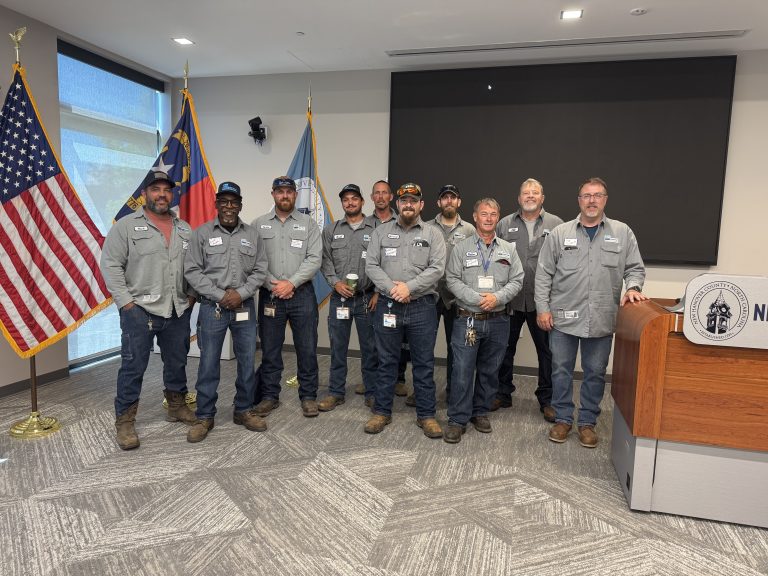 Group of utility or public works employees standing together in a meeting room near a podium and U.S. and state flags, wearing matching gray work uniforms and name badges. A large presentation screen is visible behind the group.