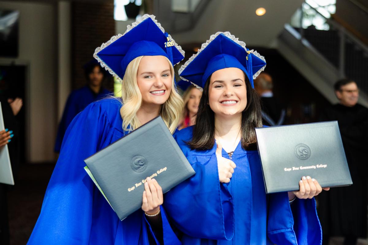 Two graduates in blue caps and gowns smiling and holding diploma covers inside a campus building during a graduation ceremony. One graduate gives a thumbs-up while both pose for the camera.