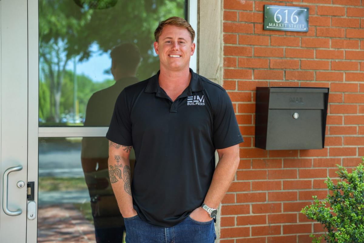 A man stands outside in front of a brick building, smiling with his hands in his pockets. He wears a black polo shirt and jeans, with a watch on his wrist and visible tattoos on one arm. A sign reading “616 Market Street” and a wall-mounted mailbox are beside him, and a glass door reflects trees in the background.
