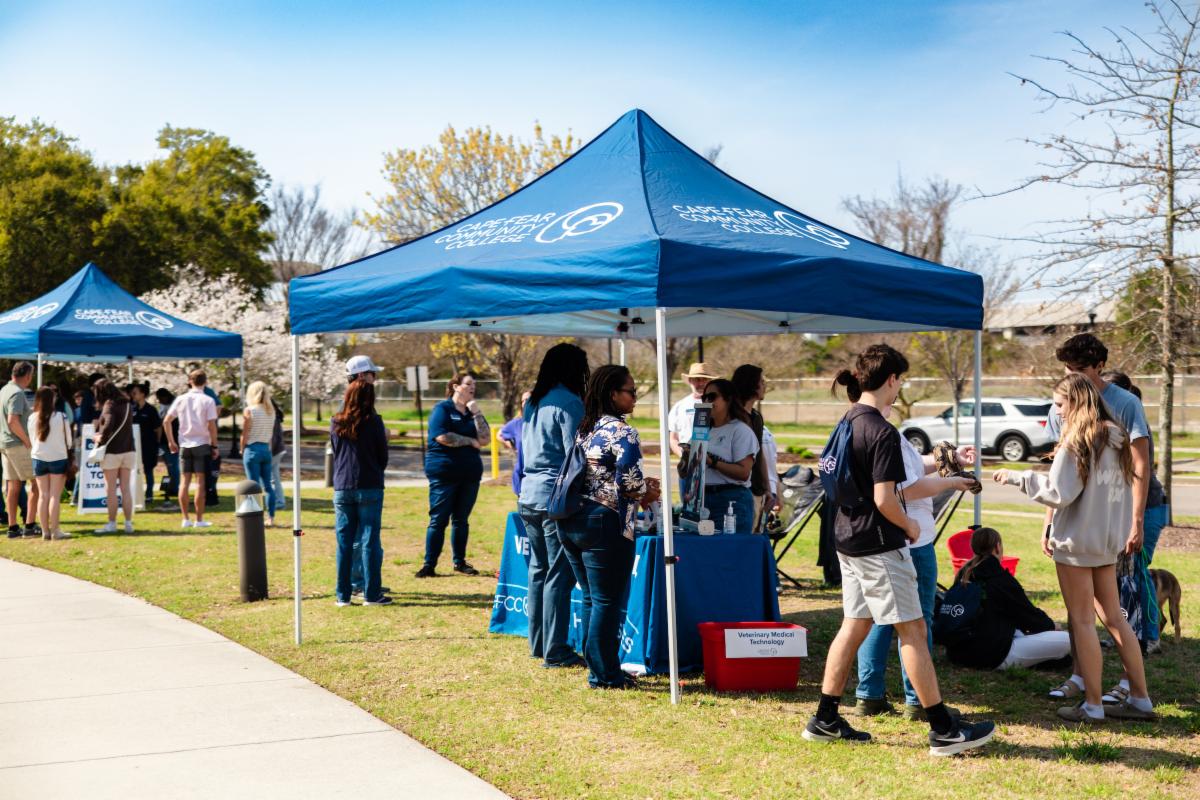 Students and staff gather under blue Cape Fear Community College tents on a grassy area, interacting at tables and informational booths during an outdoor event.