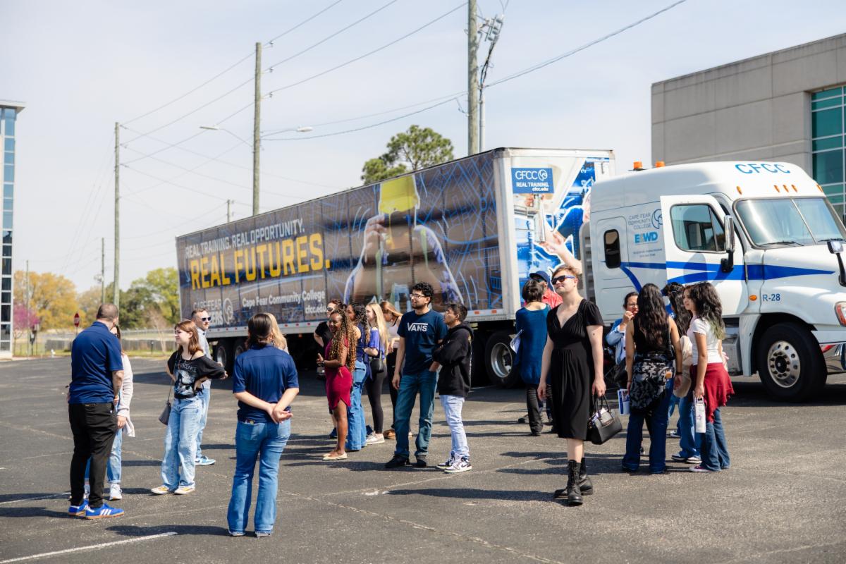 A group of people stand and talk in a parking lot beside a large Cape Fear Community College truck displaying “Real Training. Real Opportunity. Real Futures,” during an outdoor campus event.