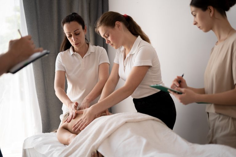 An instructor demonstrates massage techniques on a client lying on a treatment table while two students observe and take notes in a clinical training setting.