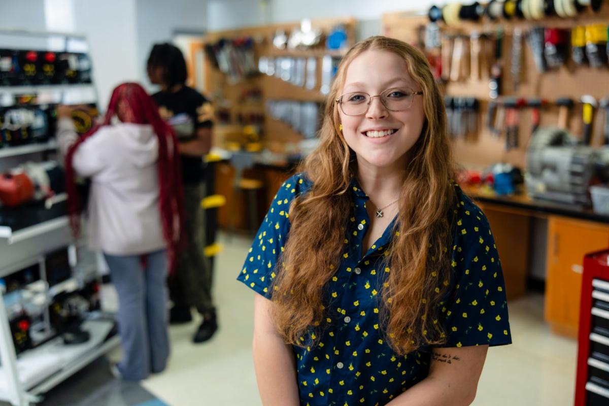 Young woman with long wavy hair and glasses smiling in a school workshop or maker space, with tools and equipment visible on the walls behind her and two students working in the background.