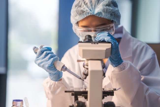 Laboratory technician wearing protective equipment, including gloves, goggles, and a hair cover, using a microscope and pipette while conducting scientific research in a lab setting.