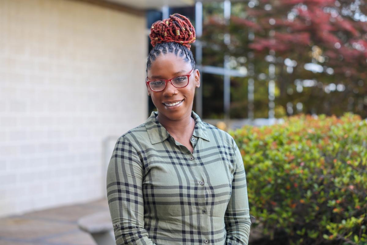 Smiling woman with braided hair in a high bun and red glasses standing outdoors near a building and landscaped bushes, wearing a green plaid button-up shirt.