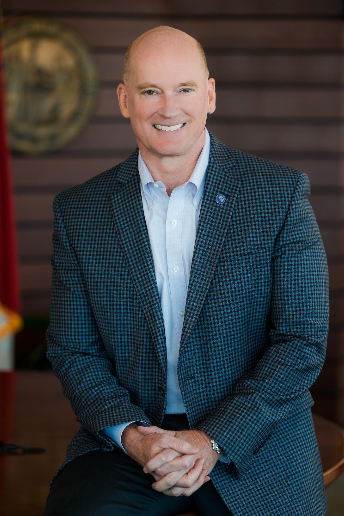 CFCC President Jim Morton sits indoors against a wood-paneled background, smiling at the camera. He is wearing a blue checkered blazer with a light blue dress shirt and a small circular lapel pin. His hands are folded in front of him as he sits on the edge of a desk, with a blurred seal and a red flag visible in the background.