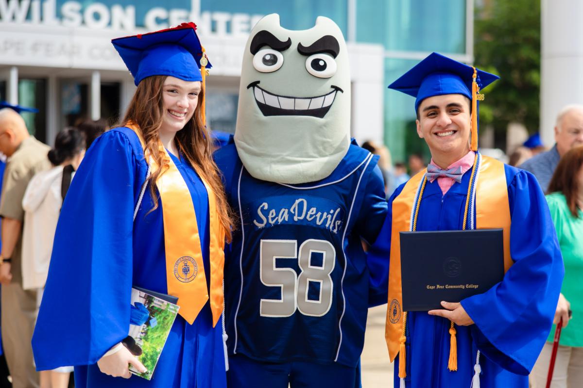 Two graduates in blue caps and gowns posing with a school mascot dressed as a “Sea Devils” character outside a campus building during a graduation ceremony. One graduate holds a diploma cover while both wear gold honor stoles and smile at the camera.