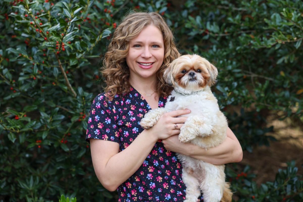 Smiling woman with curly light brown hair holds a small fluffy dog in her arms while standing outdoors in front of green foliage with red berries. She is wearing a dark top with colorful paw prints, and the dog, a small tan-and-white breed, looks calmly at the camera.