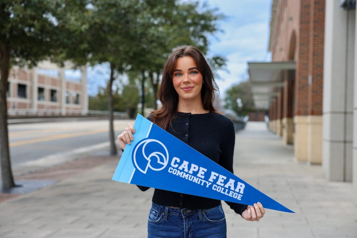 A woman stands on a sidewalk holding a blue Cape Fear Community College pennant, smiling with buildings and trees lining the street behind her.