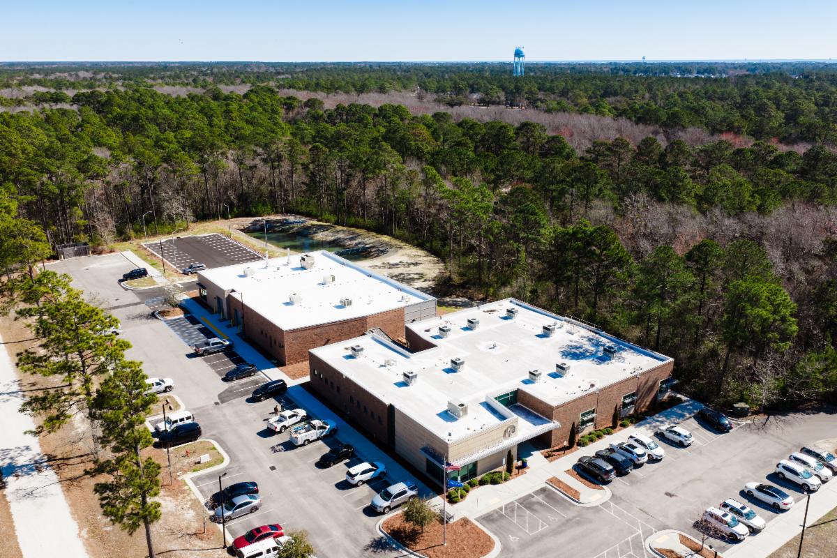 Aerial view of a brick campus building surrounded by a large parking lot and wooded area. Several cars are parked near the entrance, and a water tower is visible in the distance beyond the trees.