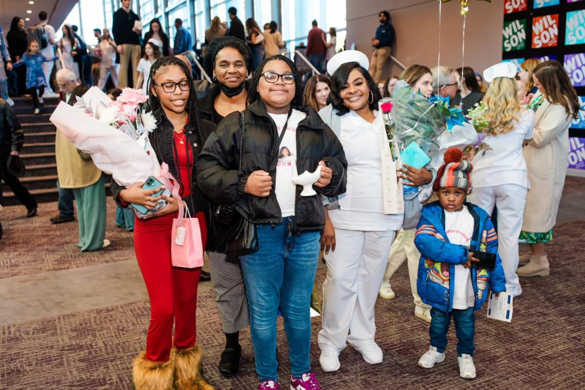 Four women and a young child pose together at a crowded indoor celebration. One woman dressed in white scrubs and a nurse’s cap holds flowers and smiles, while others hold bouquets and gifts. The group stands in a lobby with people mingling in the background, suggesting a graduation or pinning ceremony.