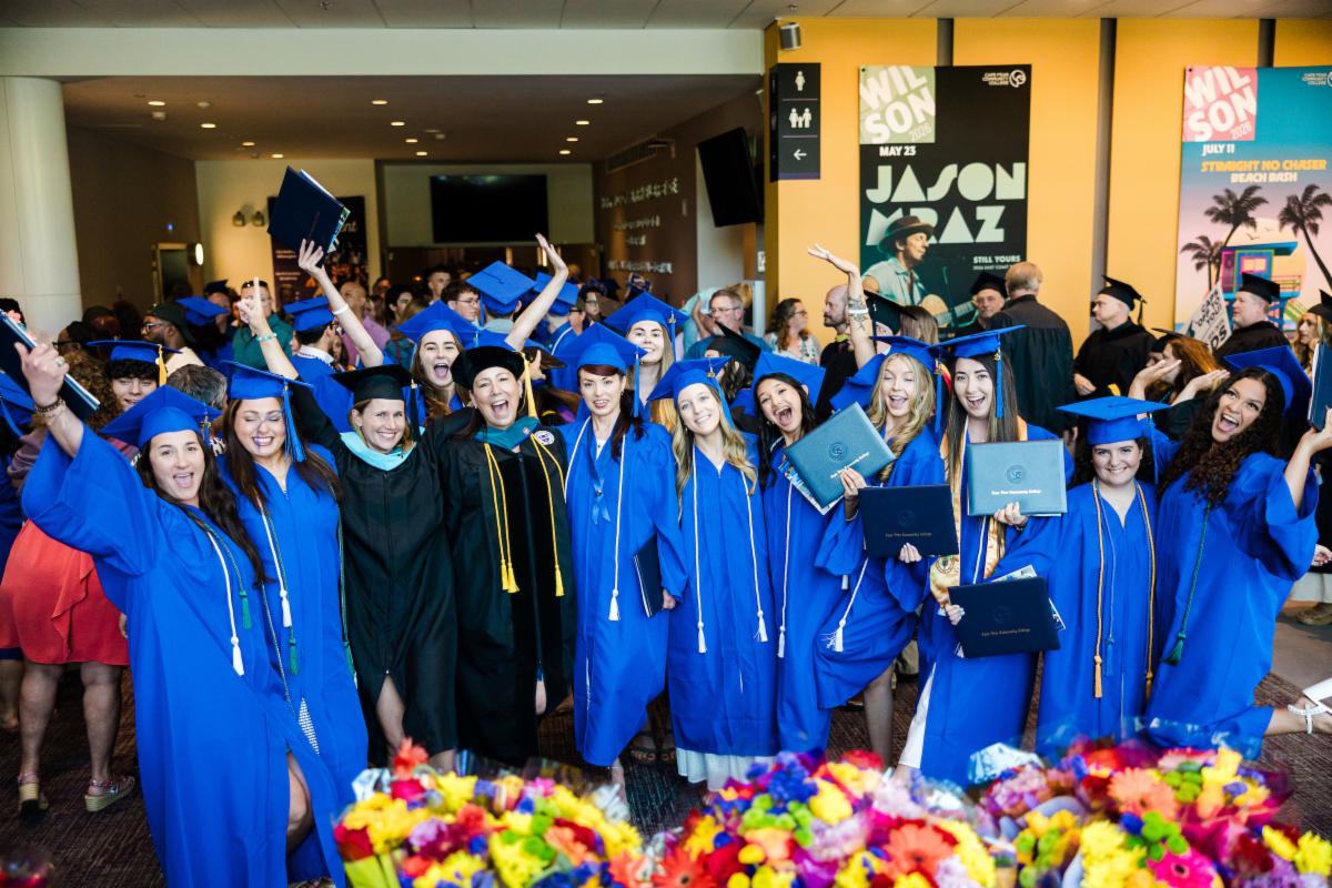 Large group of graduates and faculty members gathered inside a campus venue, smiling and celebrating with raised arms and diploma covers during a graduation event. Bright flower arrangements are visible in the foreground.