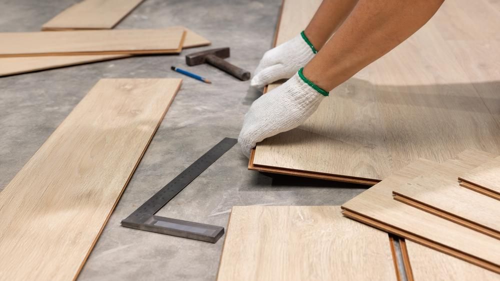 Alt text:  Close-up of a person wearing white work gloves installing light wood flooring panels on a concrete surface. Several planks are arranged nearby, along with a hammer, carpenter’s square, and pencil.