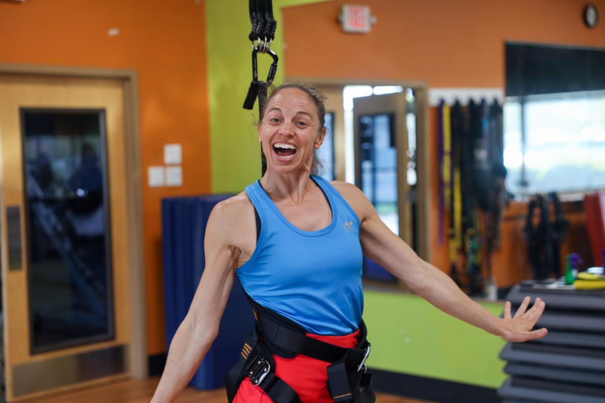 Smiling woman in a blue athletic tank top and red leggings using a suspended fitness harness in a brightly colored gym, with resistance bands and exercise equipment visible in the background.