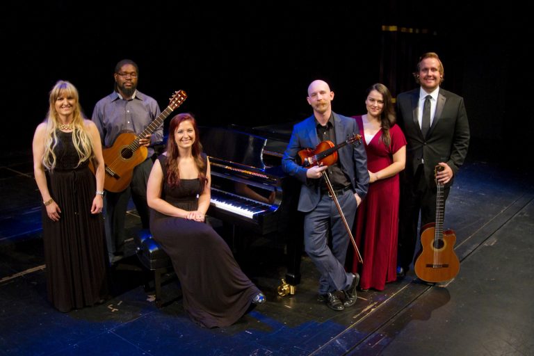 Six musicians pose on a stage around a grand piano, dressed in formal attire. Two hold guitars, one holds a violin, and one sits at the piano bench, suggesting a small ensemble or chamber group performance setting.