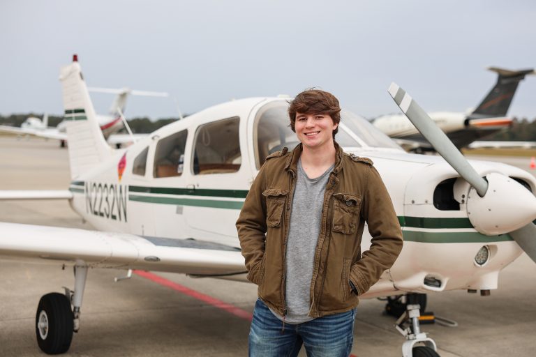 Young man standing in front of a small white propeller airplane on an airport tarmac, smiling at the camera with hands in his jacket pockets. Other aircraft are visible in the background under an overcast sky.