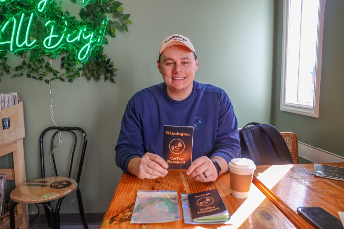 A person sits at a wooden table in a café, smiling while holding up a small booklet titled “Wilmington Coffee Passport.” They wear a light-colored baseball cap and a blue sweatshirt. On the table are additional booklets, a coffee cup with a lid, and a phone. A backpack rests on a chair to the right. Behind them, a green wall features leafy décor and a glowing neon sign, while sunlight streams in from a nearby window.
