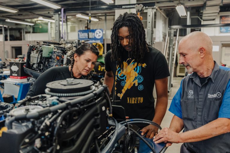 An instructor and two students examine a boat engine in a workshop, discussing components during a hands-on small marine engine training session.
