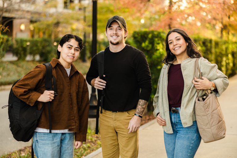 Three young adults stand outdoors on a sidewalk with backpacks, smiling at the camera. Trees with autumn foliage and greenery line the background, suggesting a campus or park setting.