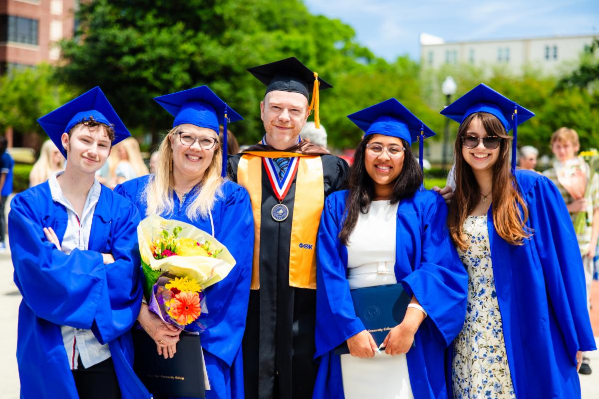 Group of graduates and a faculty member posing outdoors after a graduation ceremony. The graduates wear blue caps and gowns, and one person holds a bouquet of flowers while another holds a diploma cover.