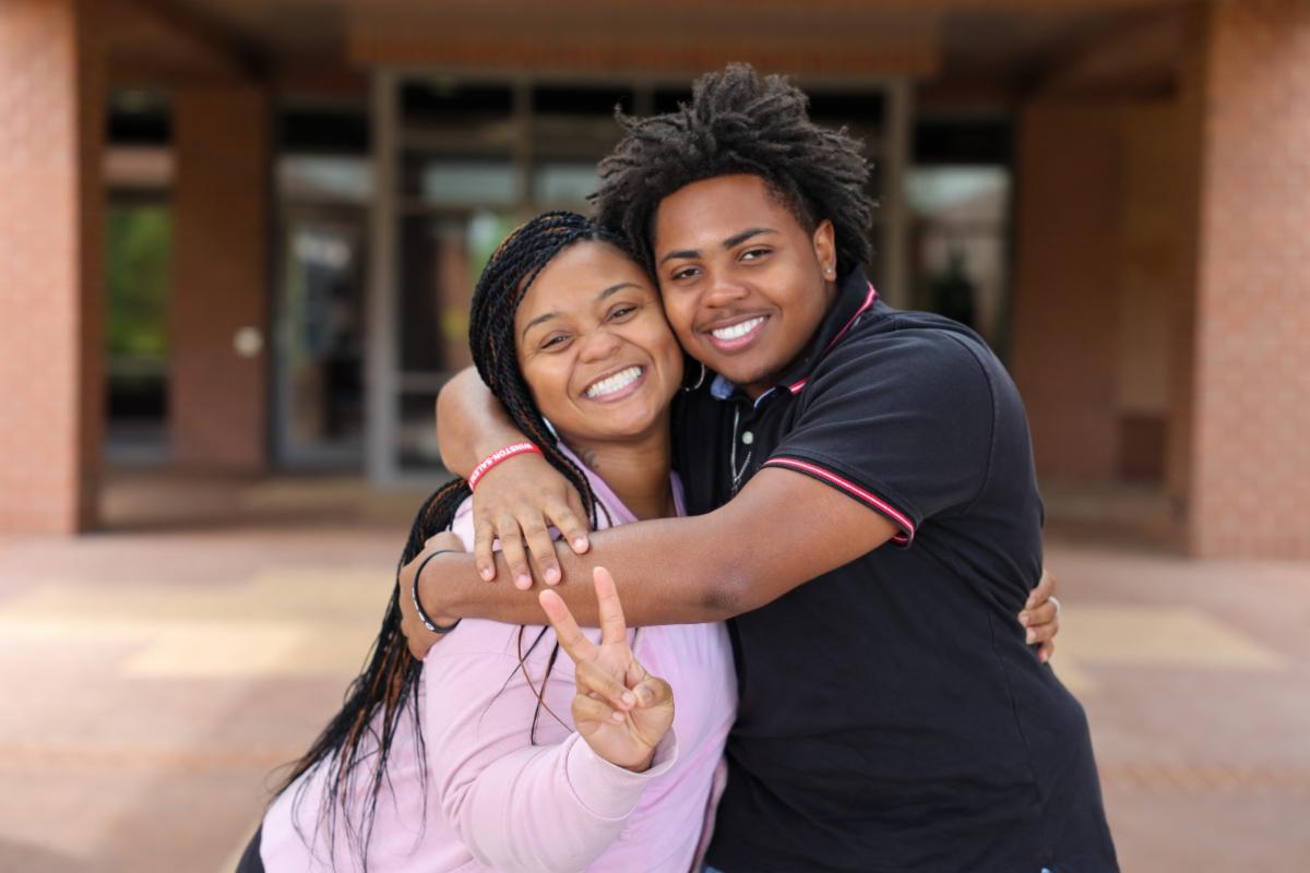 Two smiling people hugging outside a building entrance, with one person making a peace sign toward the camera.