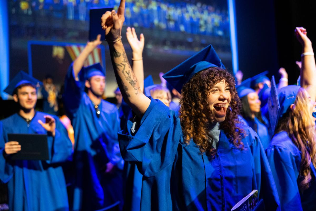 Graduate in a blue cap and gown celebrating during a graduation ceremony, smiling and raising one arm in the air while surrounded by other graduates in an auditorium.
