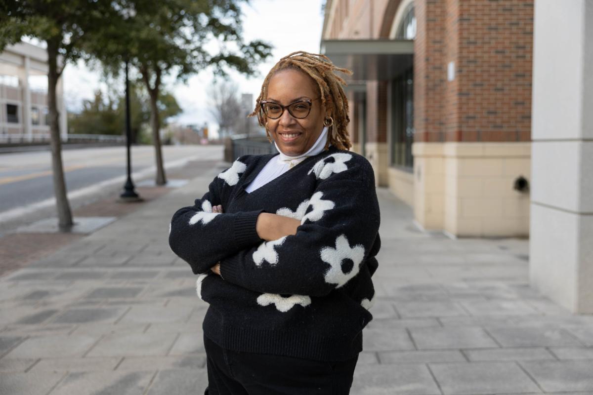 A woman wearing glasses and a black sweater with white floral patterns stands on a sidewalk with her arms crossed, smiling in front of a brick building and tree-lined street.